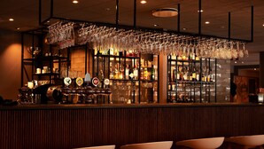 A bar area with a wooden bar counter, multiple shelves of bottles, and hanging glasses. There are three white bar stools in front. - Scandic Karlstad City (Karlstad)