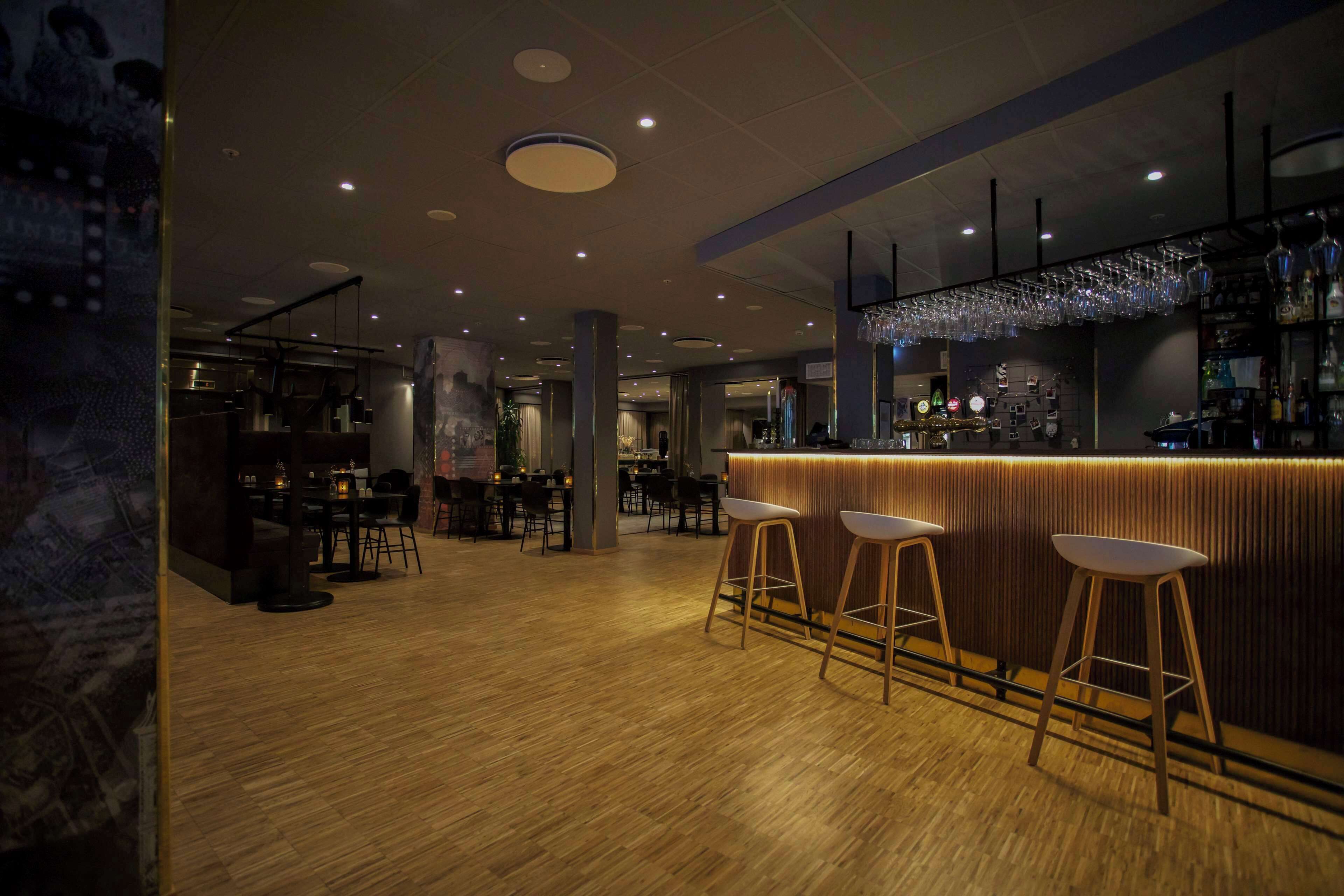 A modern bar area with wooden flooring, bar stools, and a well-lit ceiling with recessed lighting.