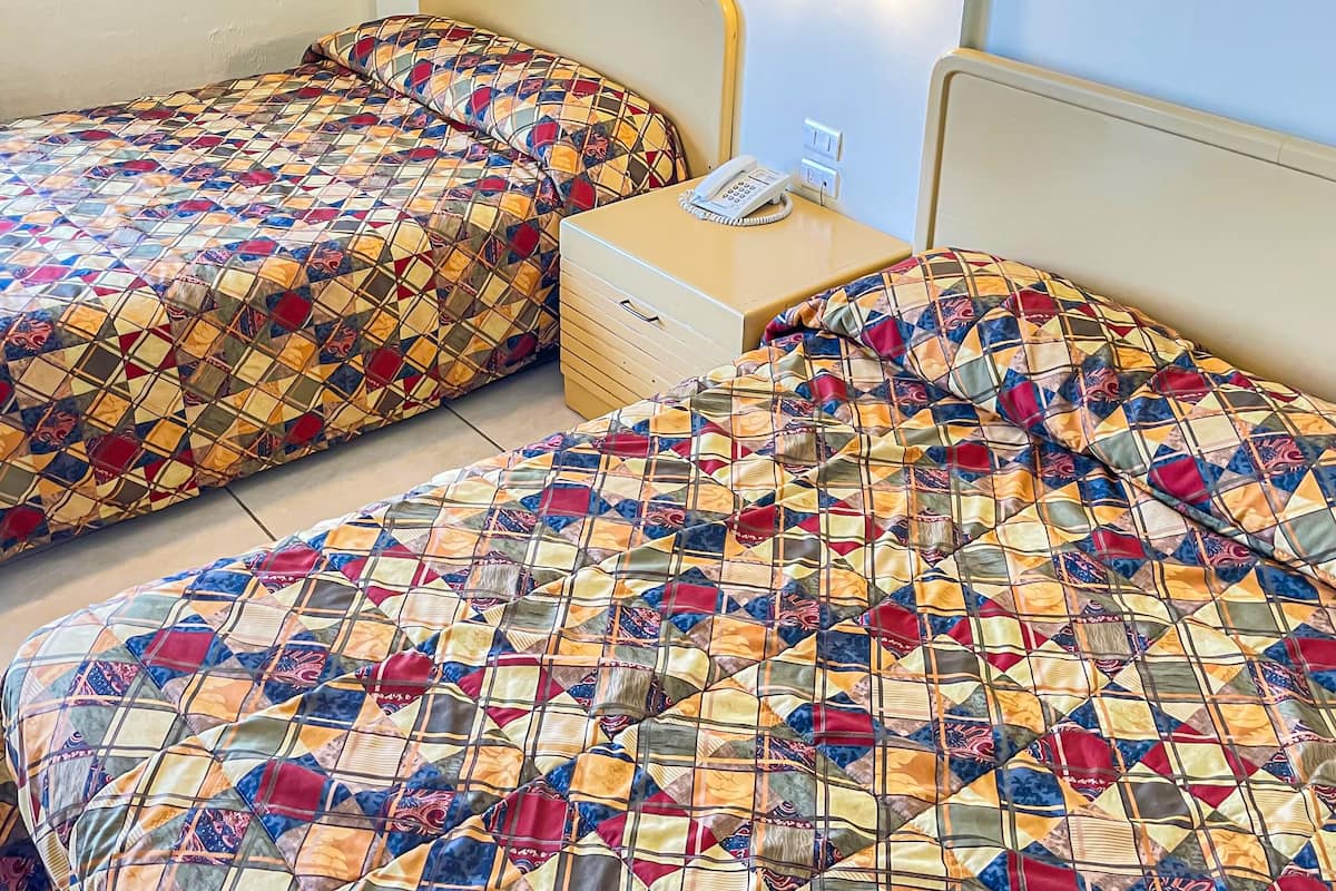 Une chambre avec deux lits, chacun recouvert d’une courtepointe à motifs. Il y a une table de chevet avec un téléphone et un interrupteur pour la lumière.