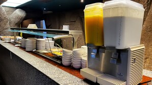 Montaje de bufé con varios dispensadores de bebidas, platos apilados y utensilios para servir.