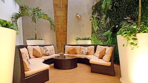 Un rincón exterior acogedor con mobiliario de mimbre y una mesa de centro de vidrio, rodeado de abundante vegetación y un muro cubierto de musgo.