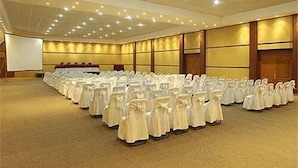 Un salón de conferencias amplio con filas de sillas blancas, una pantalla de proyector y una zona de escenario.