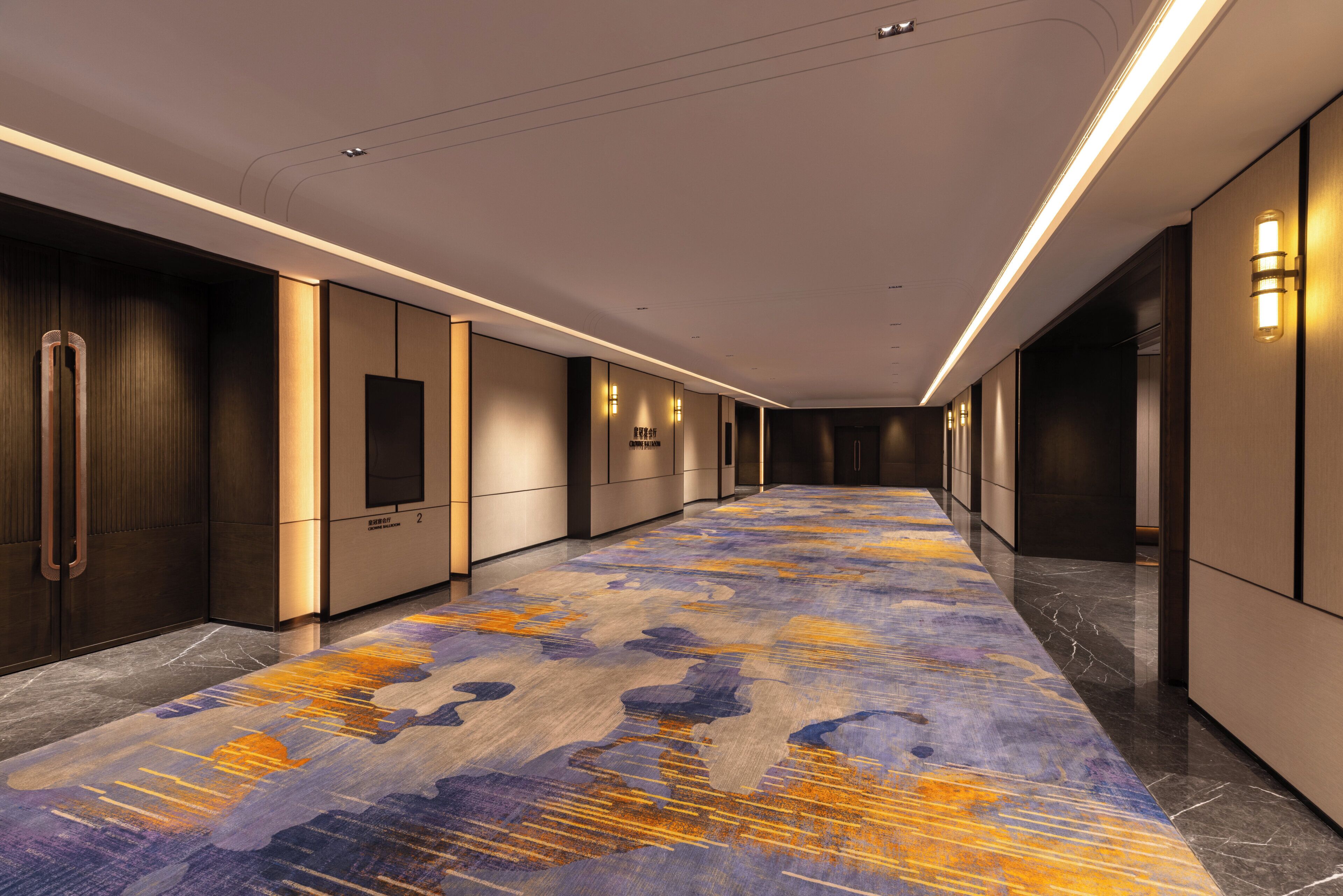 A hotel corridor with a patterned carpet, wall-mounted lights, and a modern design.