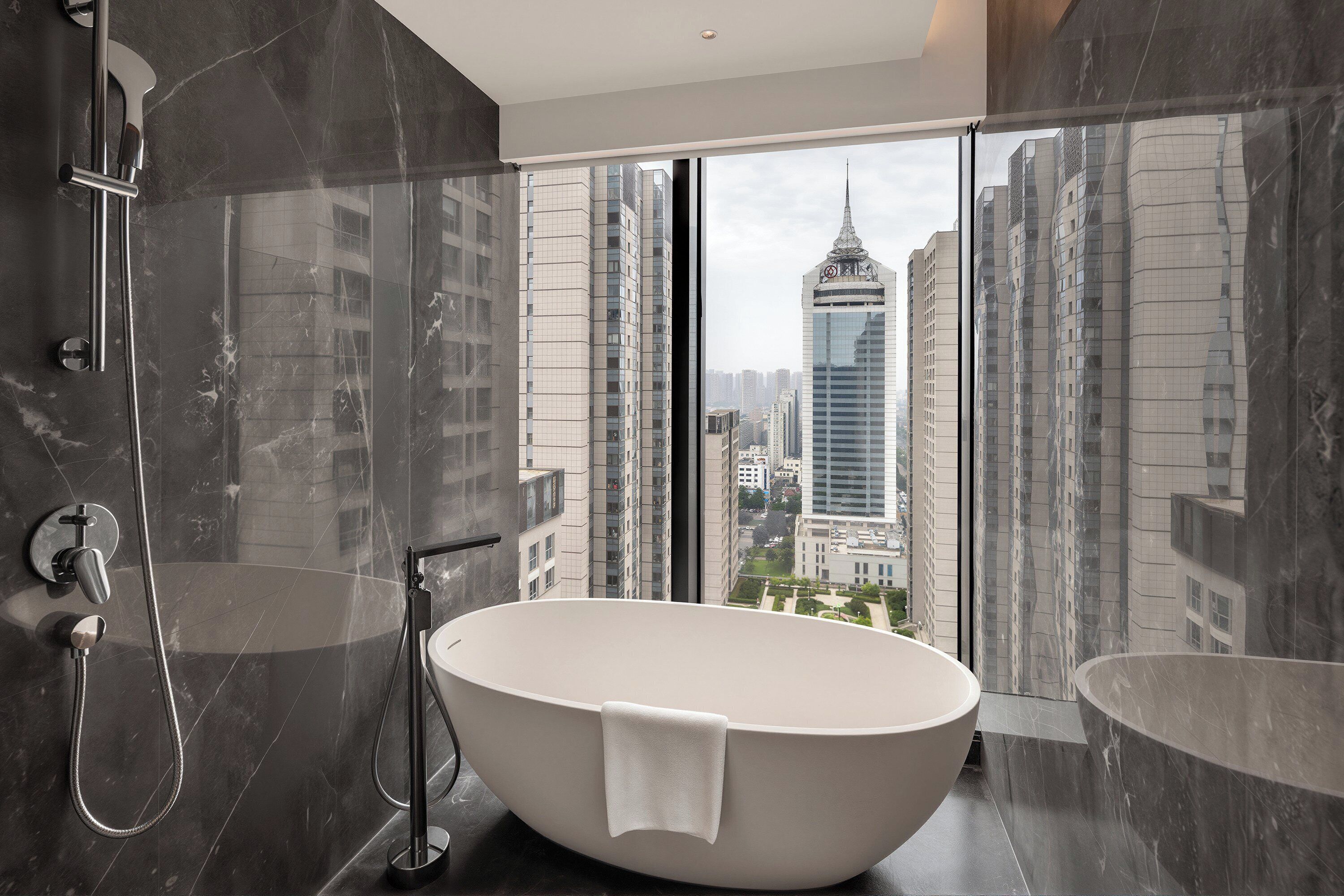 A modern bathroom with a freestanding bathtub, a glass-enclosed shower, and a view of a cityscape through large windows.