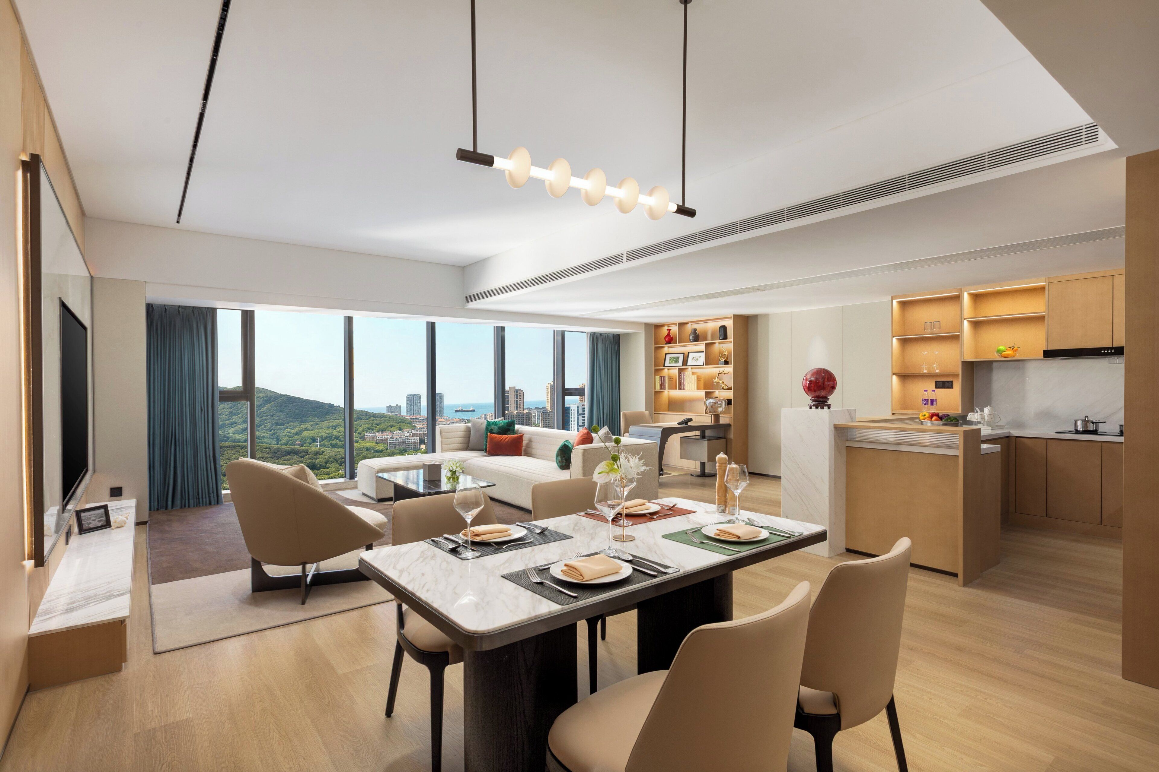 A modern hotel room with a dining area, kitchen, and a large window offering a city view.