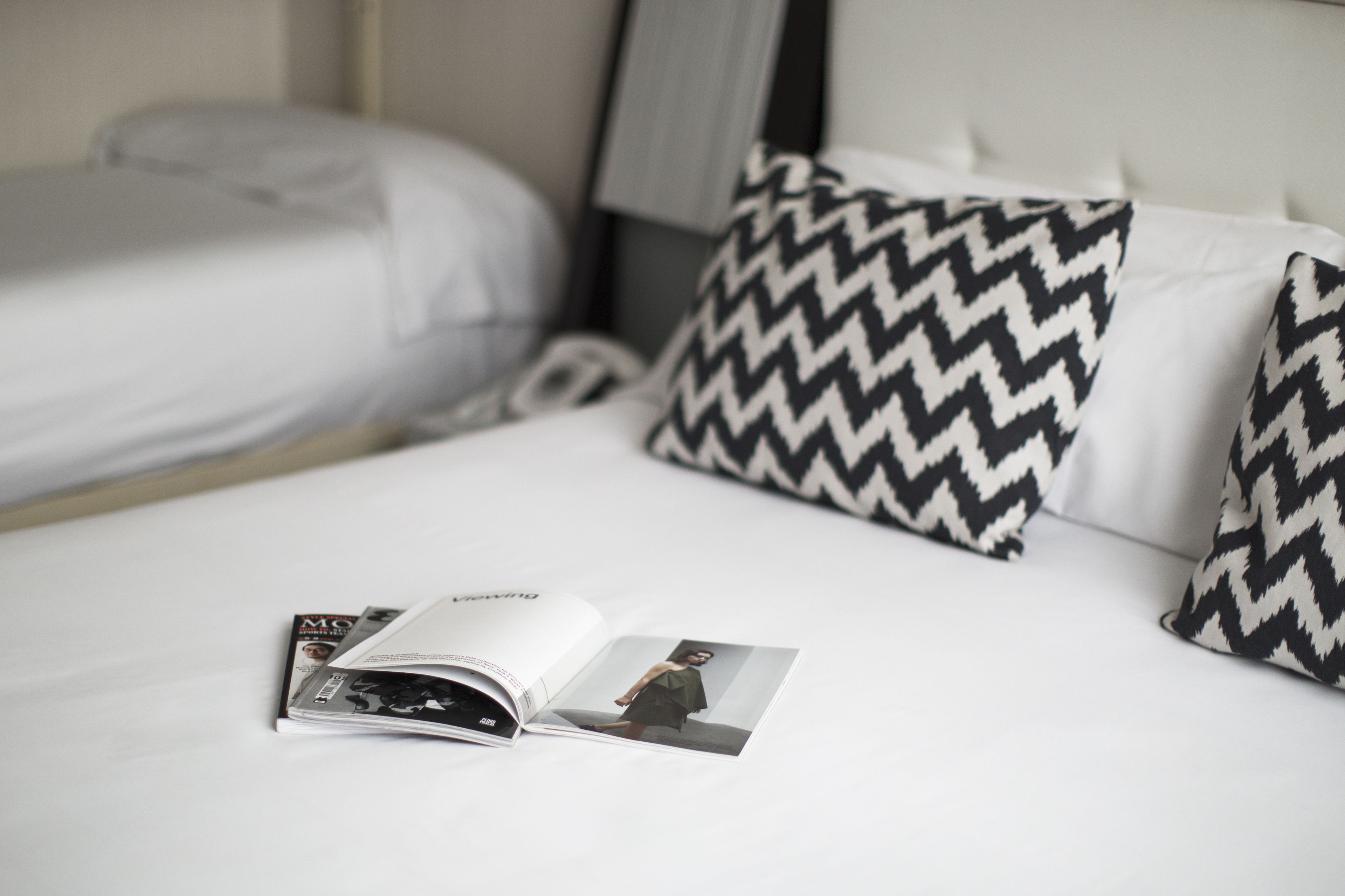 Una cama bien hecha con ropa blanca y dos almohadas estampadas, una revista sobre la cama y una mesita de noche con una lámpara.