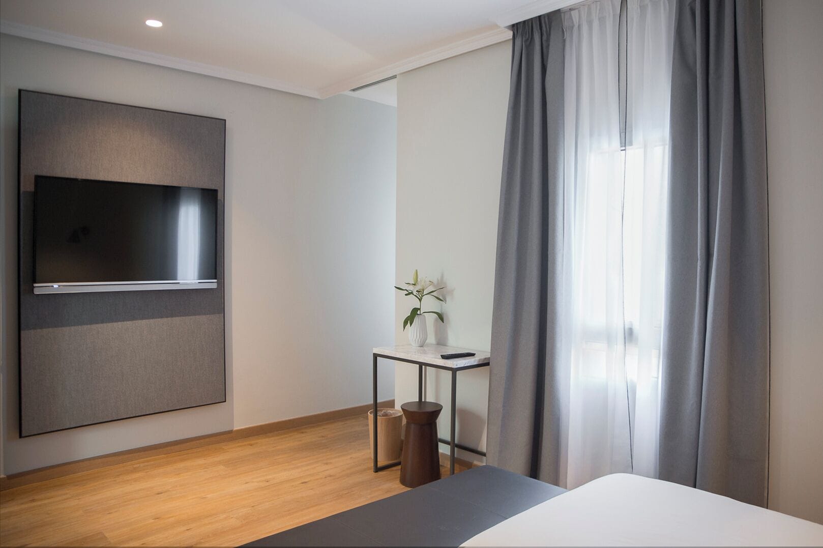 Habitación de hotel moderna con un televisor de pantalla plana grande montado en la pared, una mesita con un florero, un cesto de basura y una cama con cabecero gris.
