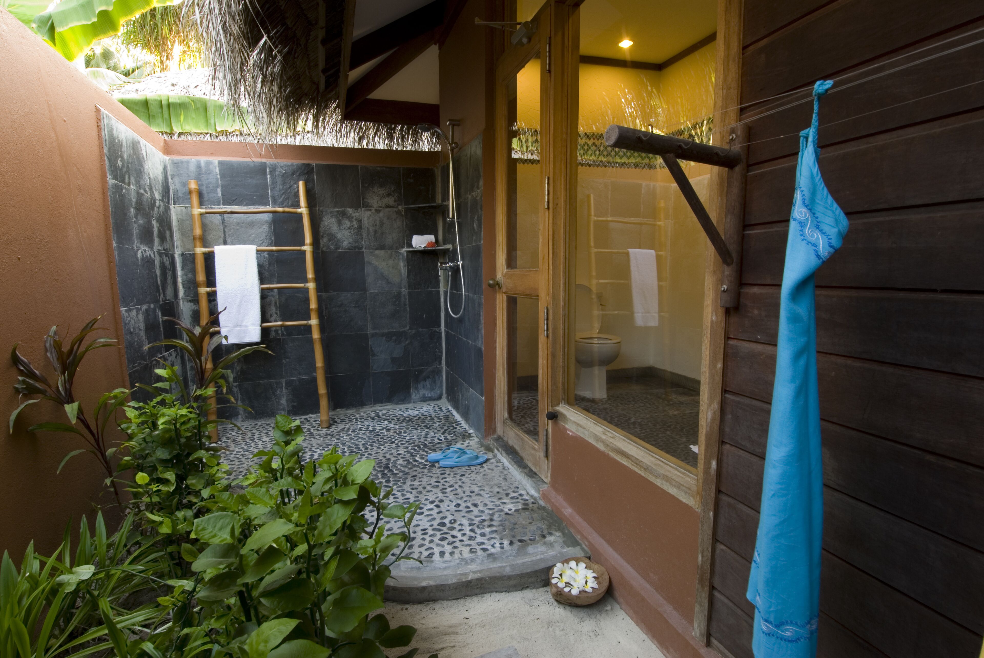 A small, private shower area with a bamboo ladder, a towel, and a blue towel hanging on a wooden wall.