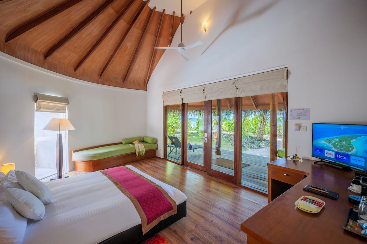 Un dormitorio con cama, escritorio, televisor y puerta corrediza de vidrio que da a un espacio exterior.