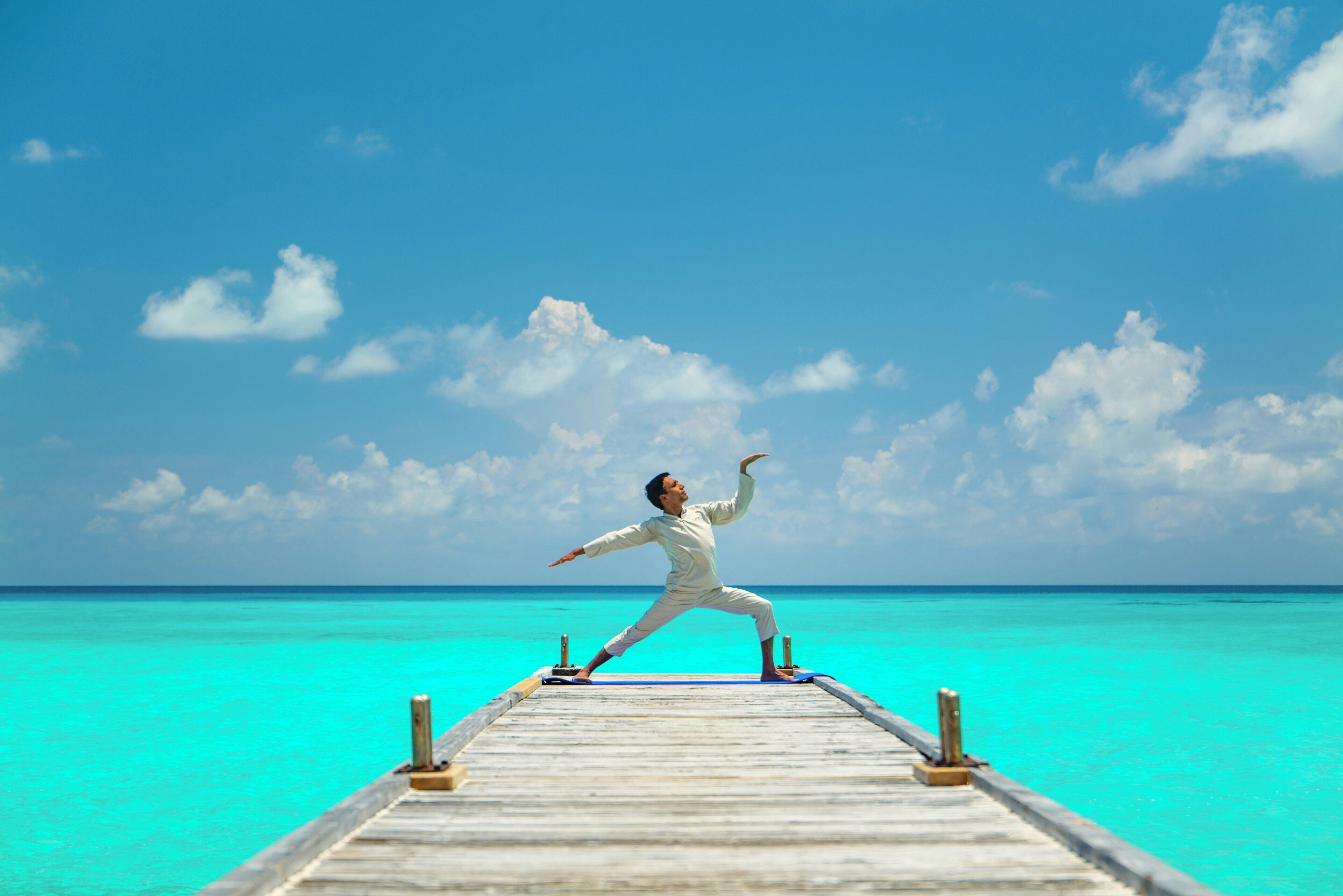 A person performing a yoga pose on a wooden pier extending into a turquoise sea under a clear sky.