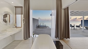 A modern bathroom with a bathtub, a round mirror, and a view of a deck with outdoor seating. - Niva Kuramathi Maldives (Kuramathi Island)
