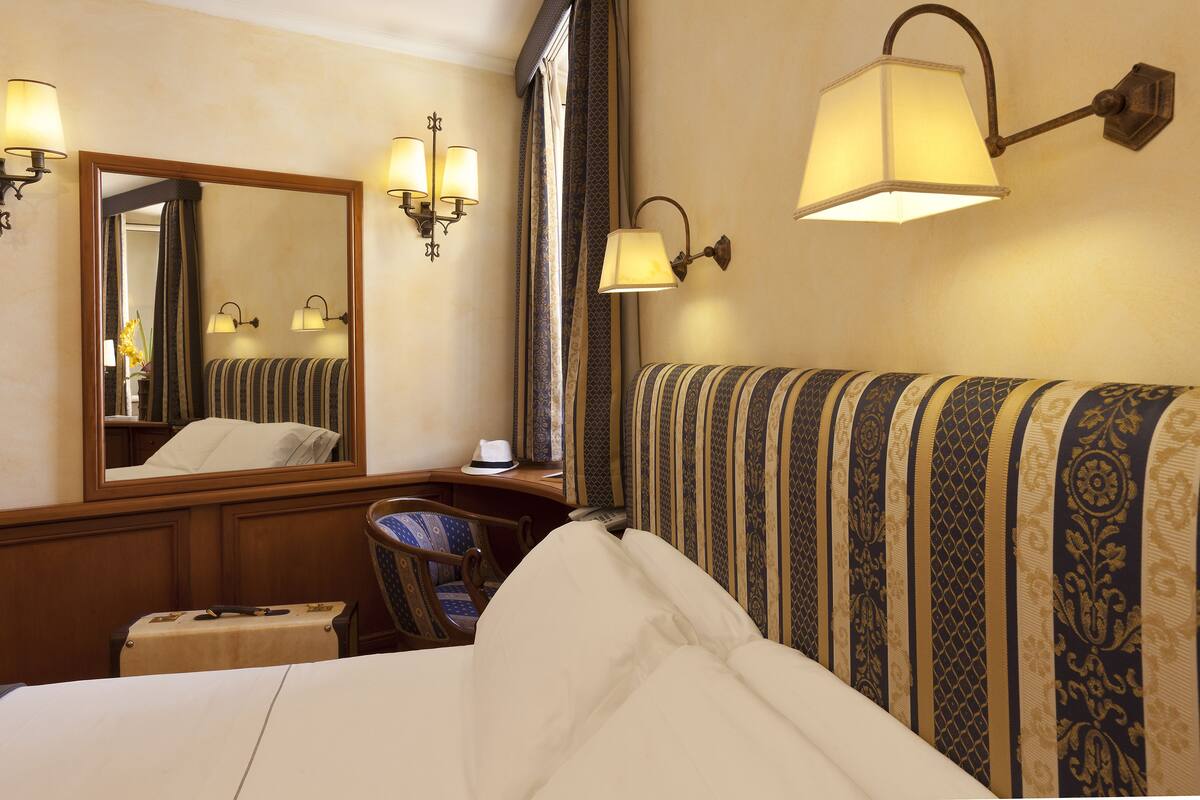 A hotel room with a large bed, a chair, a desk, and a mirror. There are wall sconces with lamps and a window with curtains.