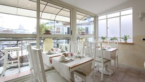 A dining area with white furniture, large windows, and potted plants. - La Fenice (Rome)