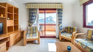 A living room with a television, wooden furniture, and a view of mountains through a large window. - Apartamentos Coral Beach (Calpe)