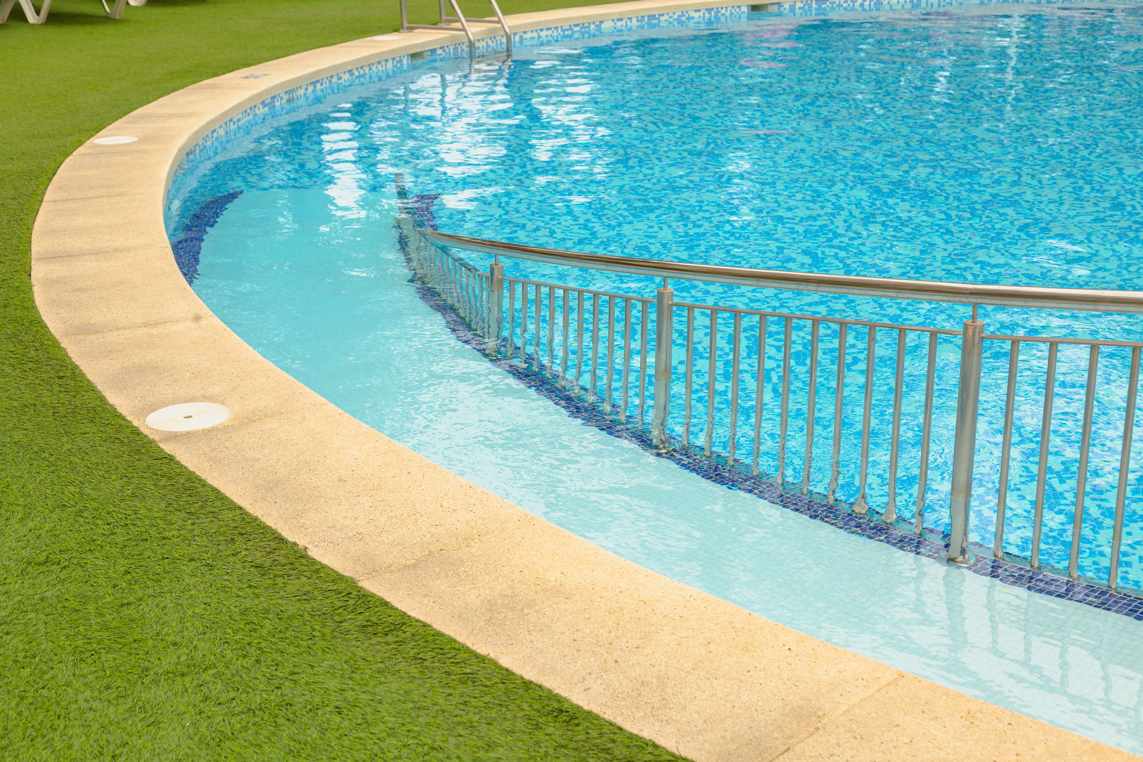 Une piscine avec un bord carrelé, une rambarde de sécurité en métal et une zone herbeuse qui l’entoure.