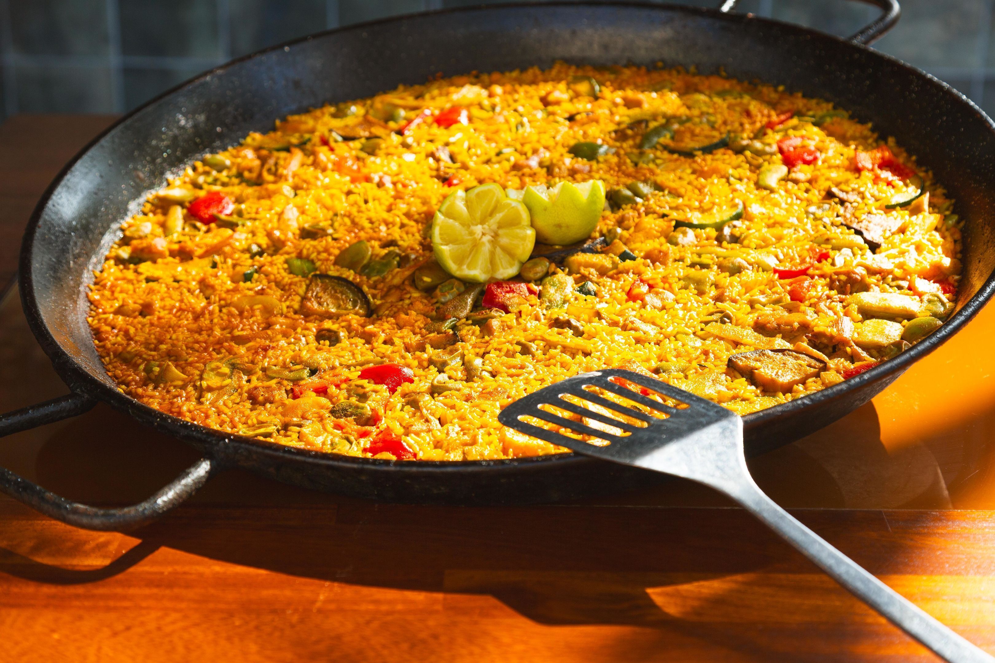 Una sartén grande y poco profunda, llena de un plato de arroz amarillo, adornado con rodajas de lima y una espátula negra.