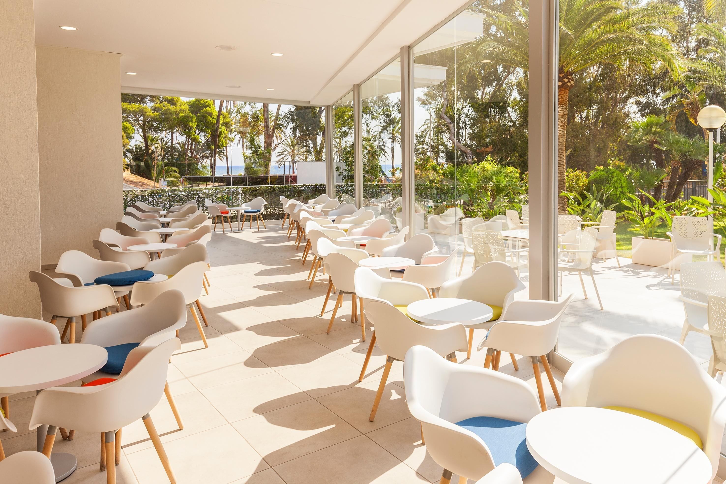 A modern outdoor seating area with white chairs and tables, large windows, and a view of palm trees and greenery.