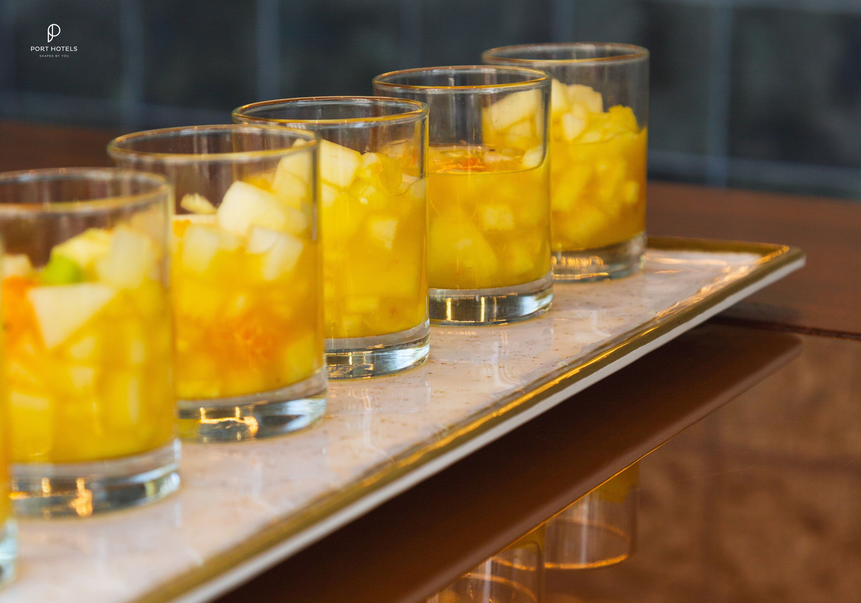 A tray with six glasses containing a yellow beverage and diced fruit.