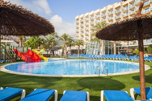 A swimming pool with blue lounge chairs and a slide, surrounded by palm trees and a hotel building. - Port Denia Hotel (Denia)