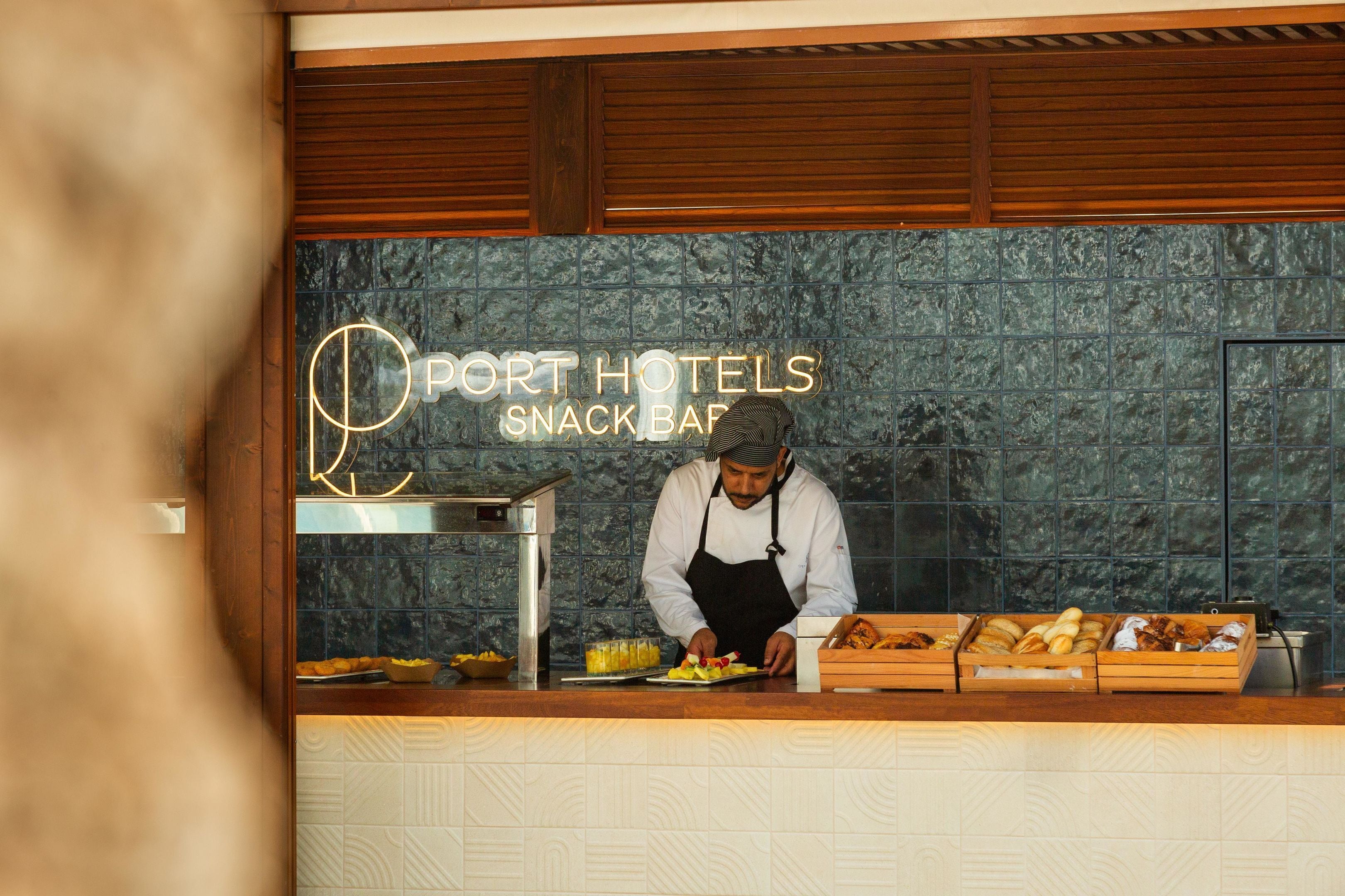 Ein Koch bereitet Essen an einer Theke zu, auf der ein Schild mit der Aufschrift "Port Hotels Snack Bar" steht.