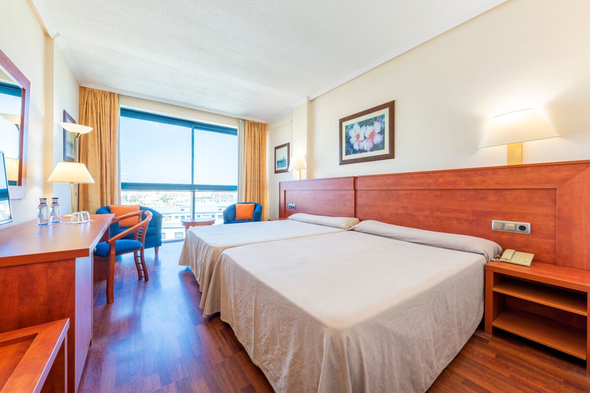A hotel room with two beds, a desk, a chair, a lamp, and a window with a view.