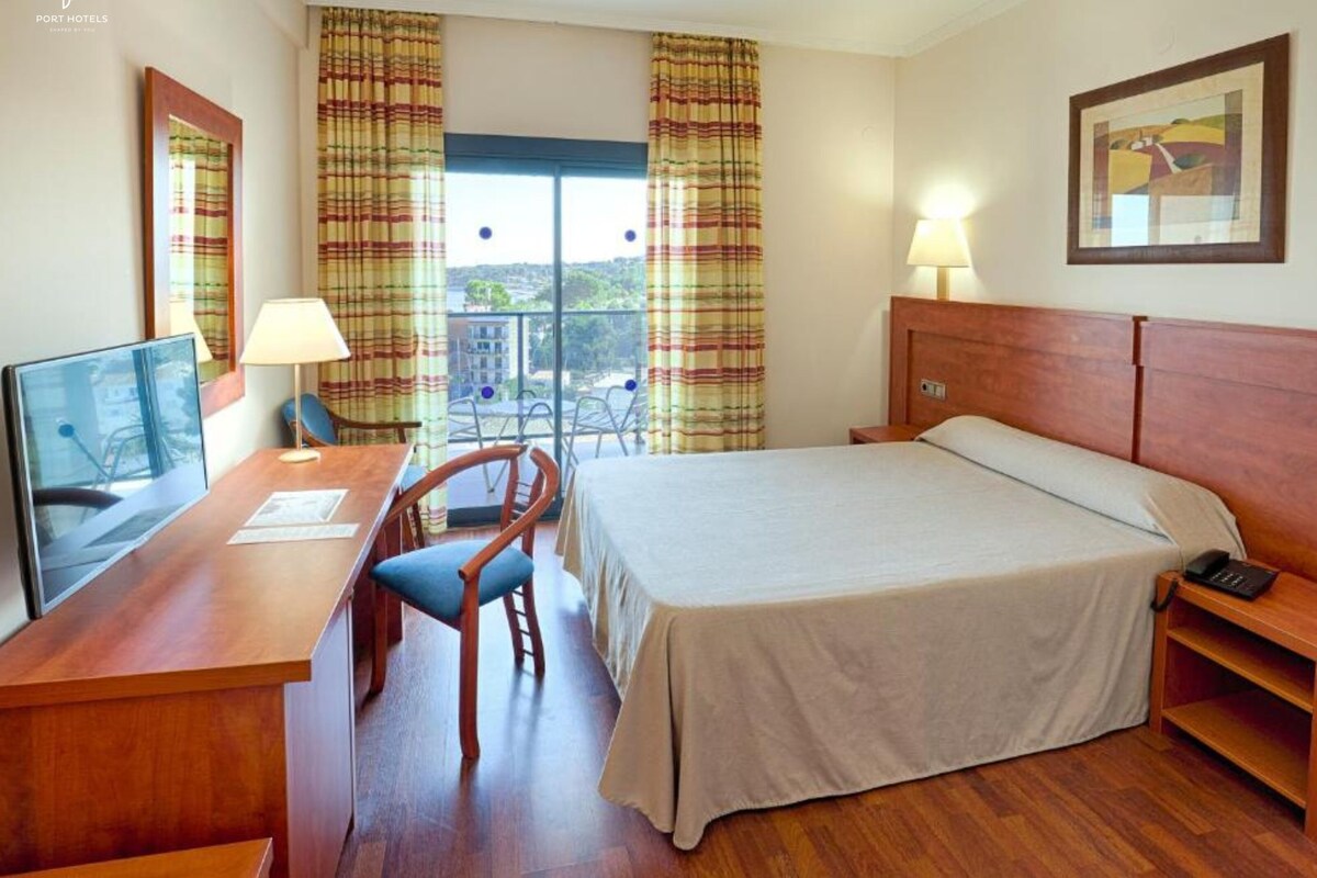 A hotel room with a bed, desk, chair, and a view of the city through large windows.