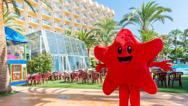 A red mascot standing in a resort area with palm trees, outdoor seating, and a modern building in the background.