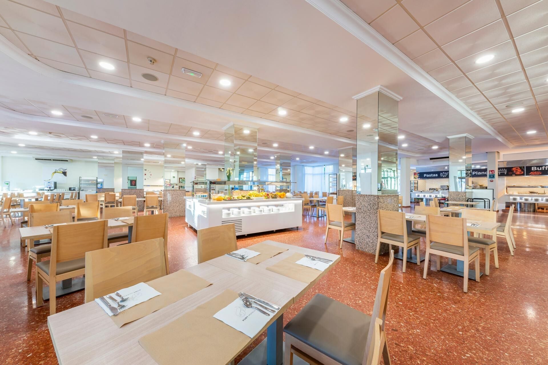 Una cafetería amplia con varias mesas y sillas, un mostrador de comida central, y un interior luminoso y bien iluminado.