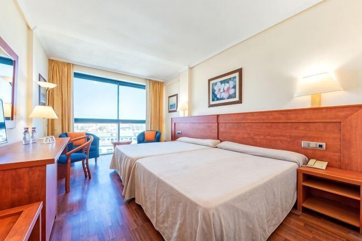 Habitación de hotel con dos camas, un escritorio, una silla, ventana con vistas y un cuadro en la pared.