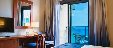 A hotel room with a bed, a desk with a television, a chair, a lamp, and a balcony with a view of the city.