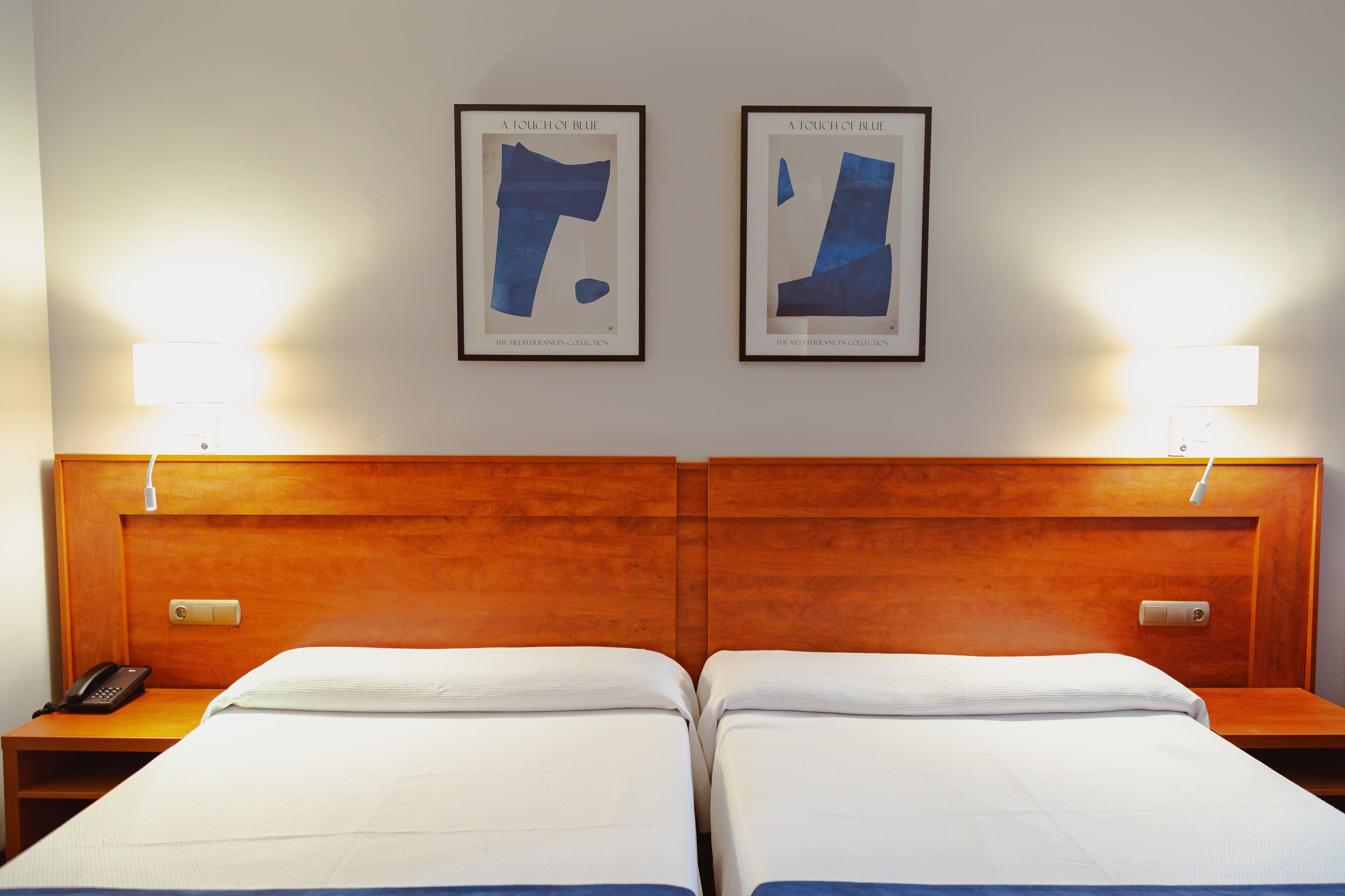 A hotel room with a wooden headboard, two beds with white bedding, a bedside table with a phone, and two framed artworks titled "A Touch of Blue" on the wall.