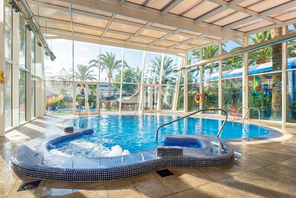 An indoor swimming pool with a hot tub, clear glass walls, and a view of palm trees and a playground. - Port Denia Hotel (Denia)
