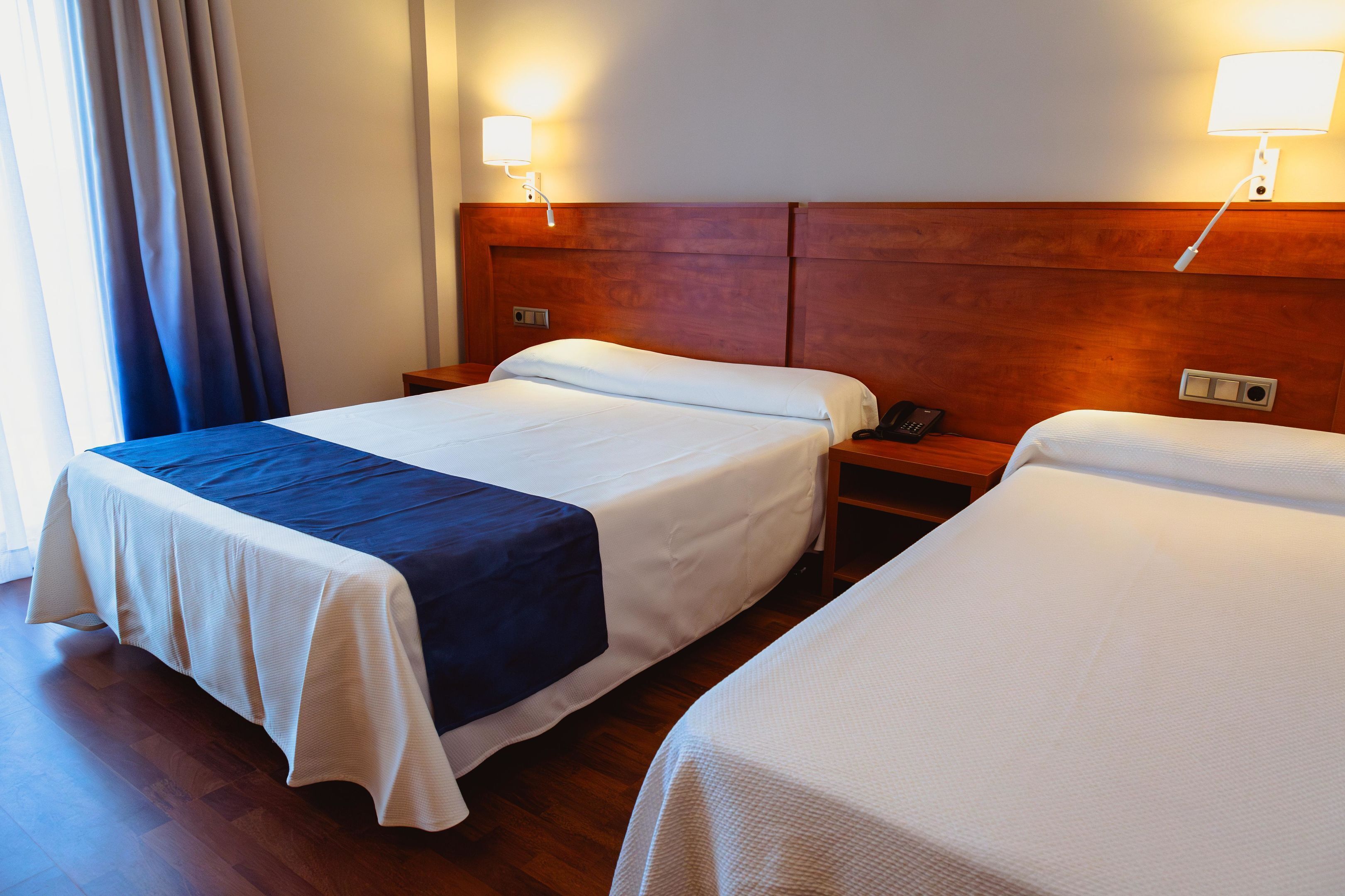 A hotel room with two beds, wooden headboards, bedside lamps, a telephone, and a window with curtains.