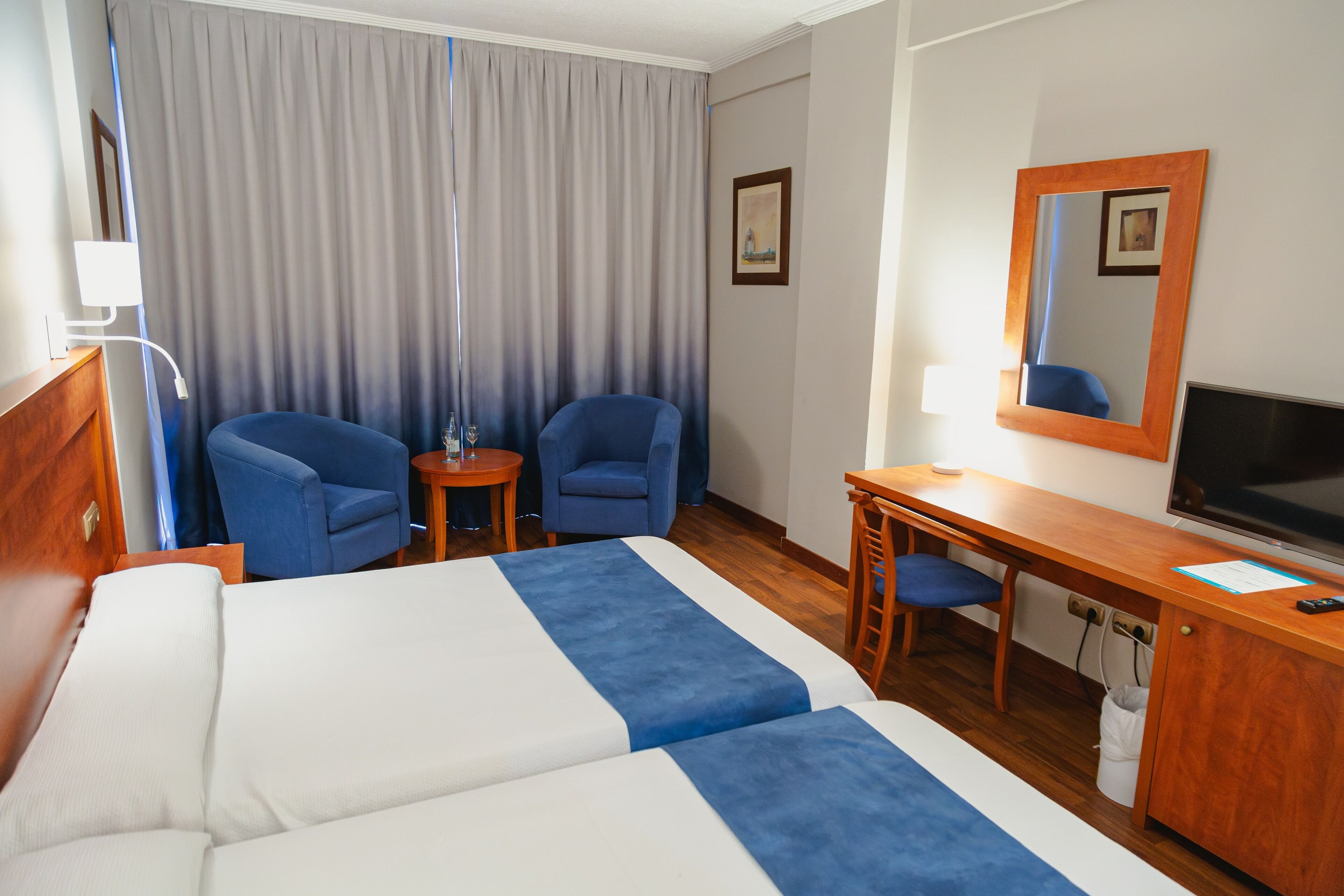 Ein Hotelzimmer mit zwei Betten, einem Schreibtisch, einem Sessel, einem Fernseher und einem Fenster mit Vorhängen.