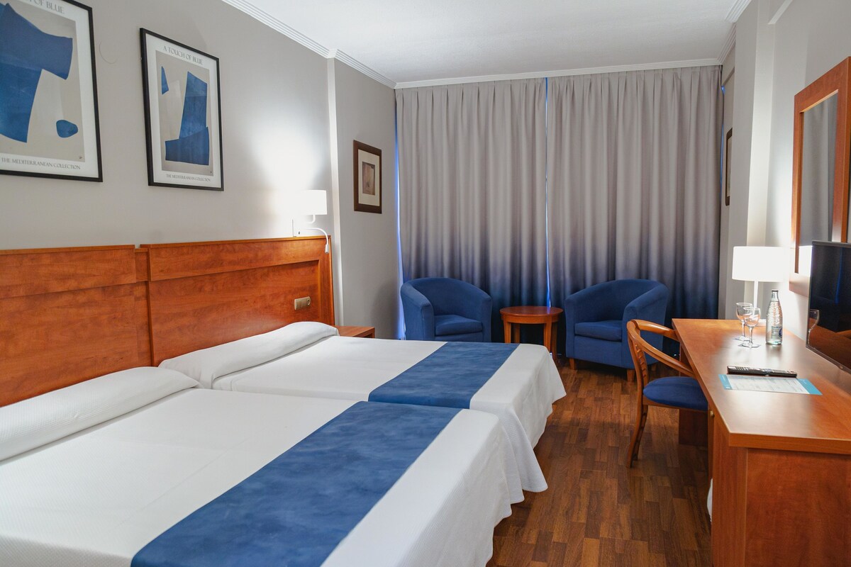 A hotel room with two beds, a desk with a chair, a small table, and a large window with curtains.