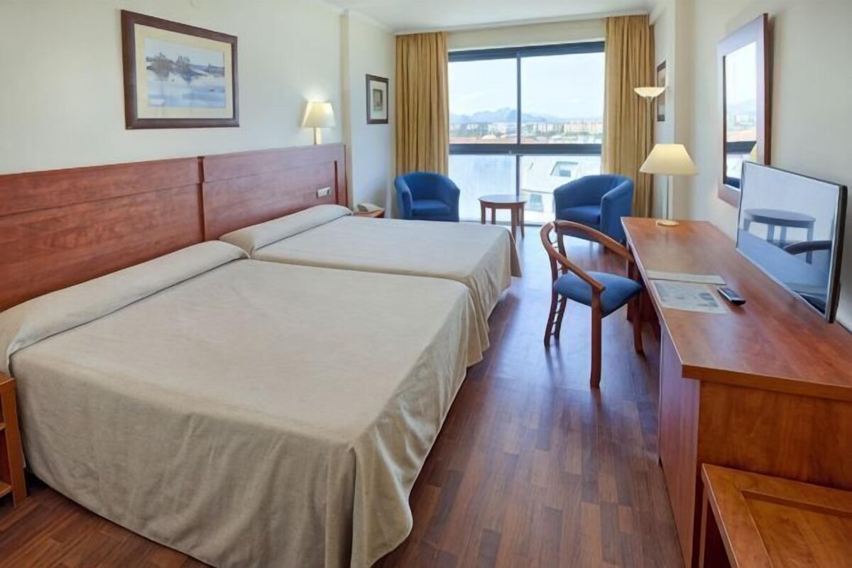 Habitación de hotel con dos camas, un escritorio, una silla, un televisor y una ventana con vistas.