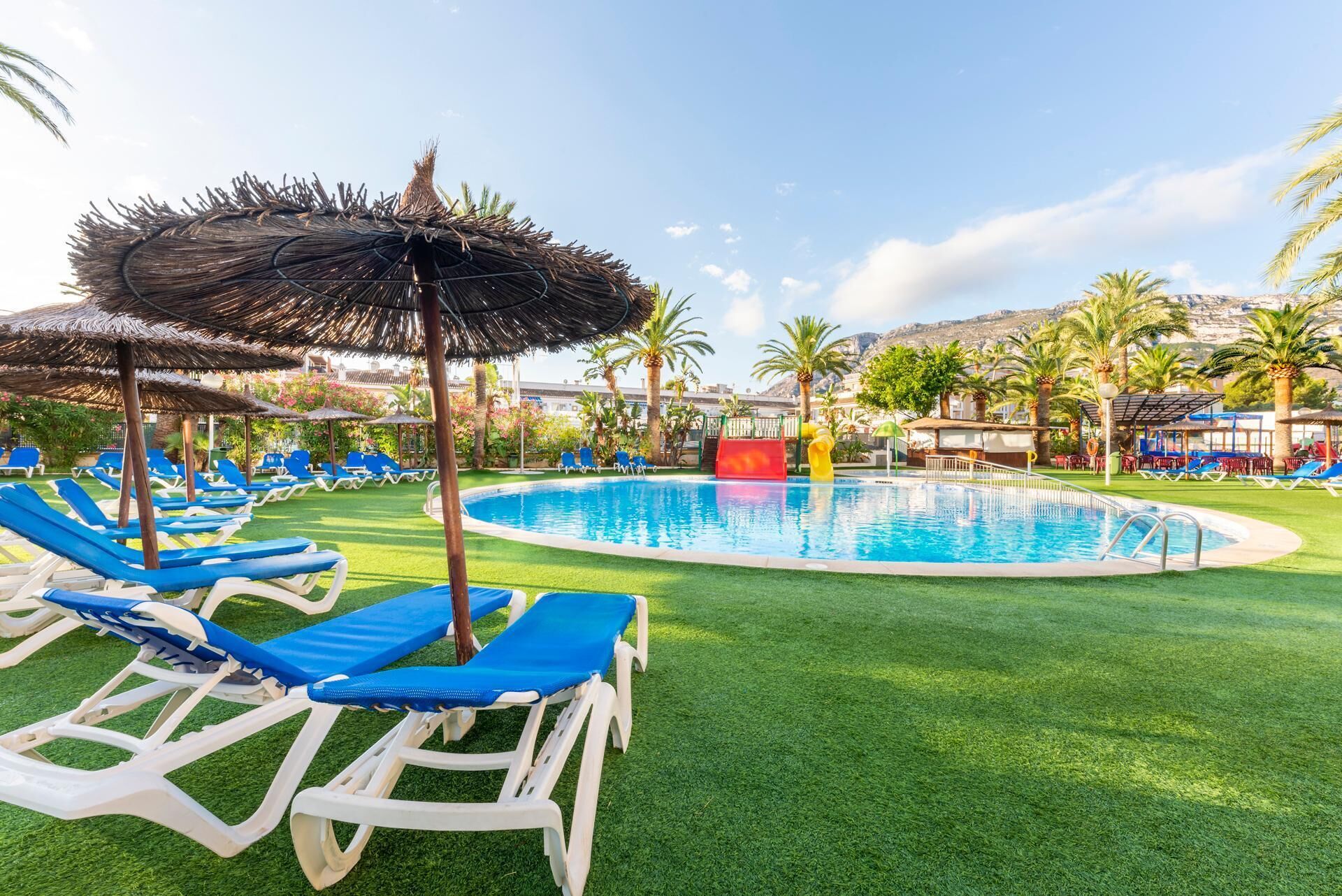 Zona de piscina con sillas reclinables azules, una sombrilla de paja y un tobogán rojo.