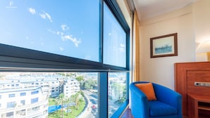 A modern hotel room with a large window offering a city view, a blue armchair, and a wooden side table. - Port Denia Hotel (Denia)