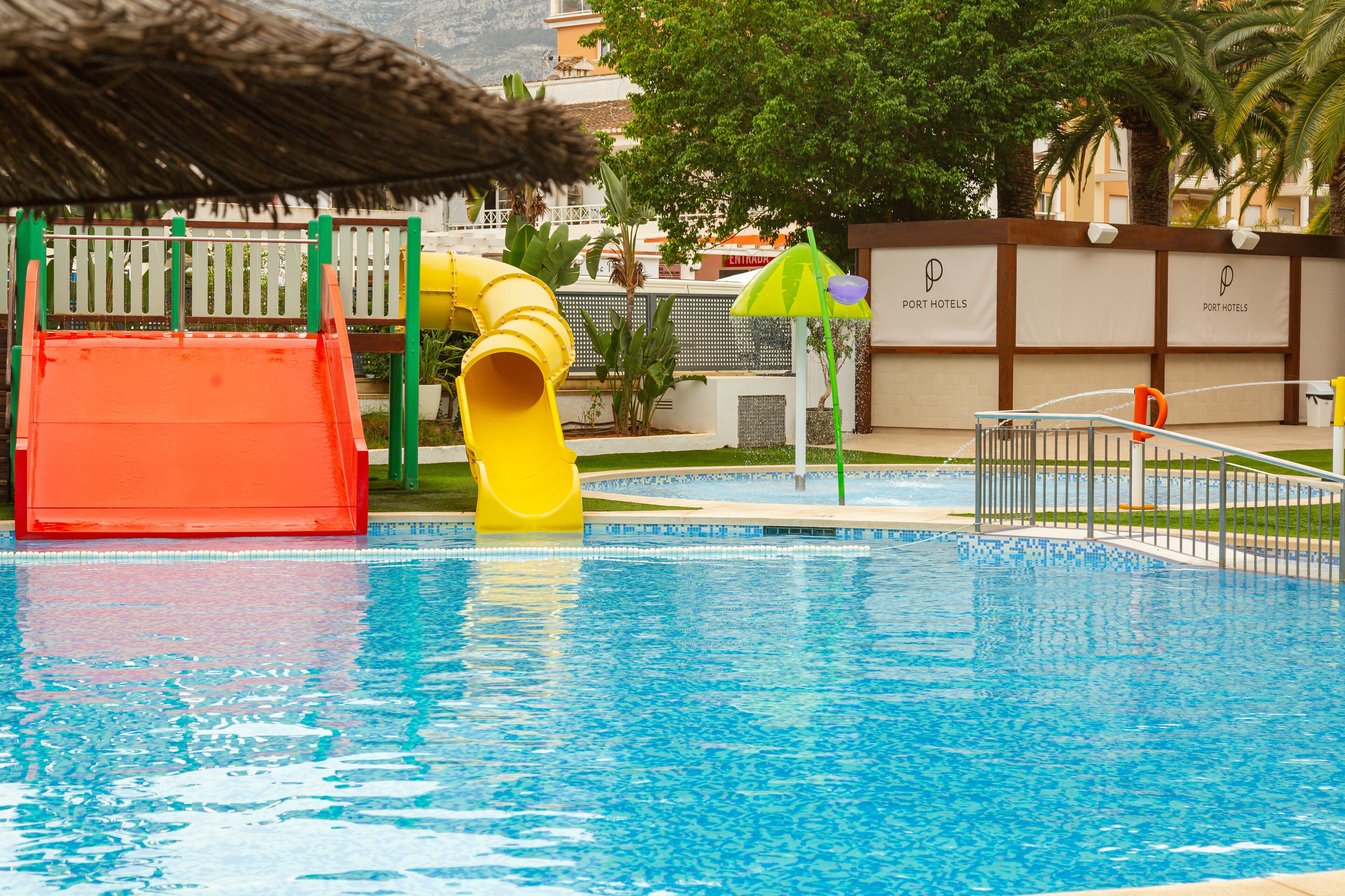 A swimming pool with a slide, a thatched umbrella, and a fence with signage.