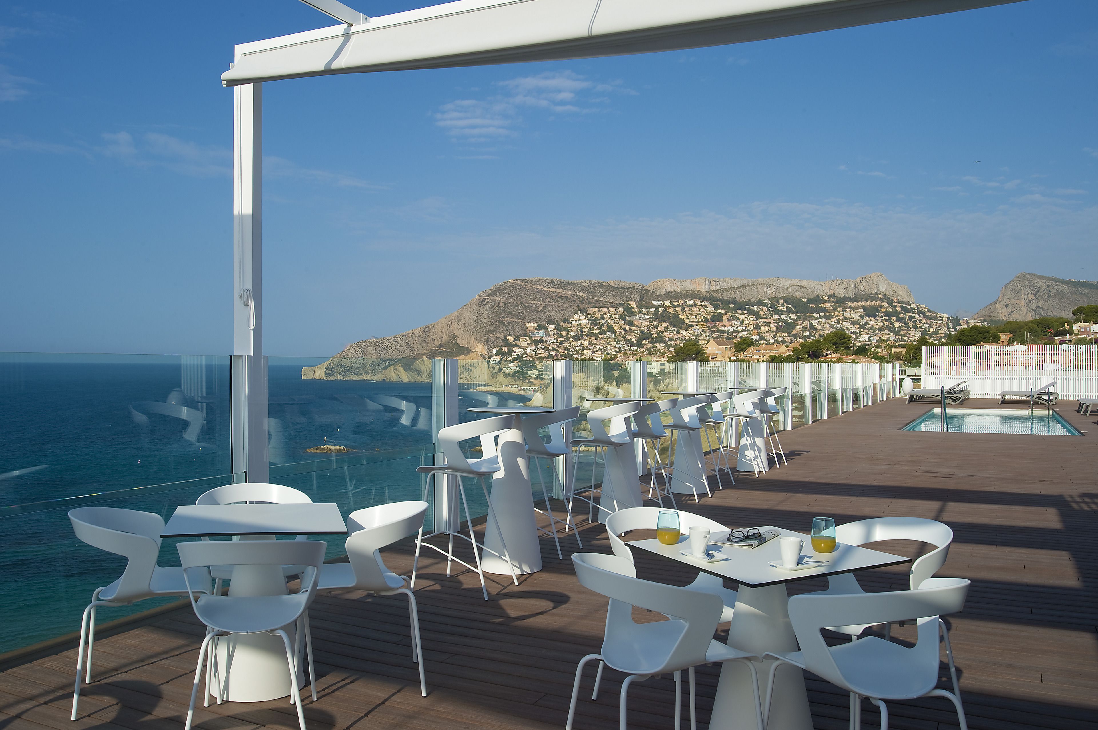 Une terrasse avec des chaises et des tables blanches, donnant sur une piscine et un paysage montagneux.
