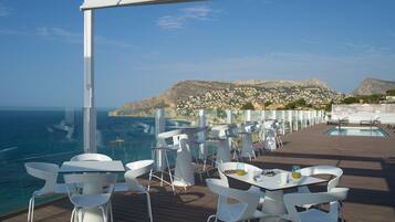 Une terrasse avec des chaises et des tables blanches, donnant sur une piscine et un paysage montagneux.