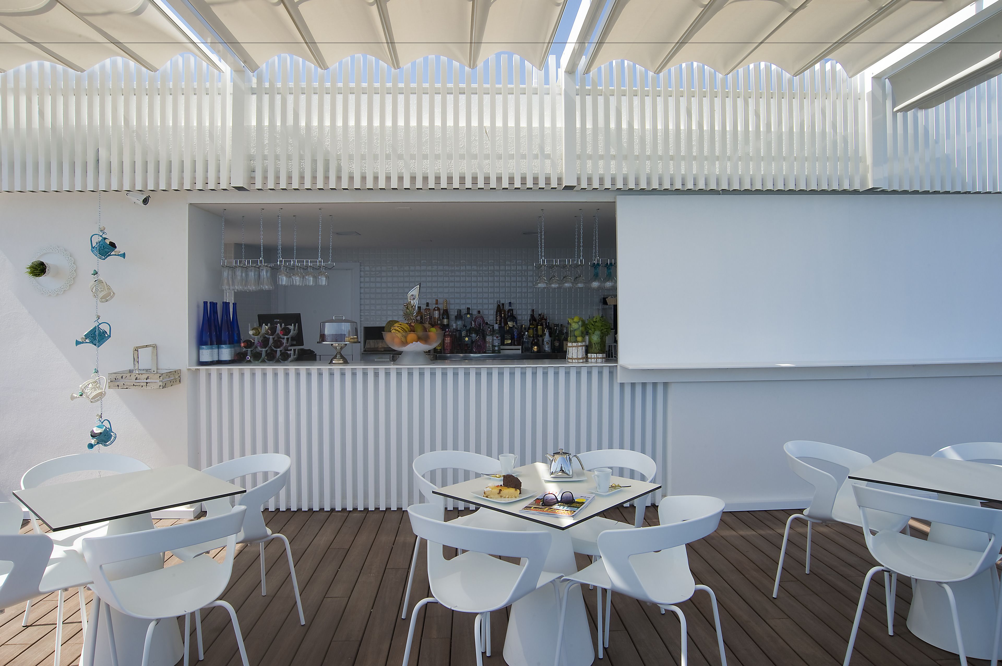 Un área de bar moderna al aire libre con sillas y mesas blancas, una barra con diferentes botellas y una valla decorativa blanca.