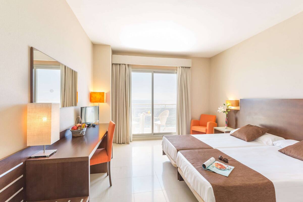 A hotel room with a bed, desk, and chair, a large window with curtains, and a view of the city.