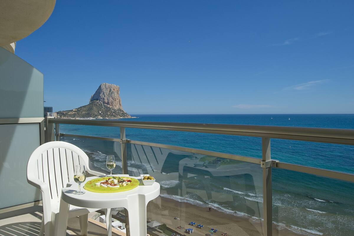 A balcony with a table set for two, overlooking a beach and a rocky cliff.