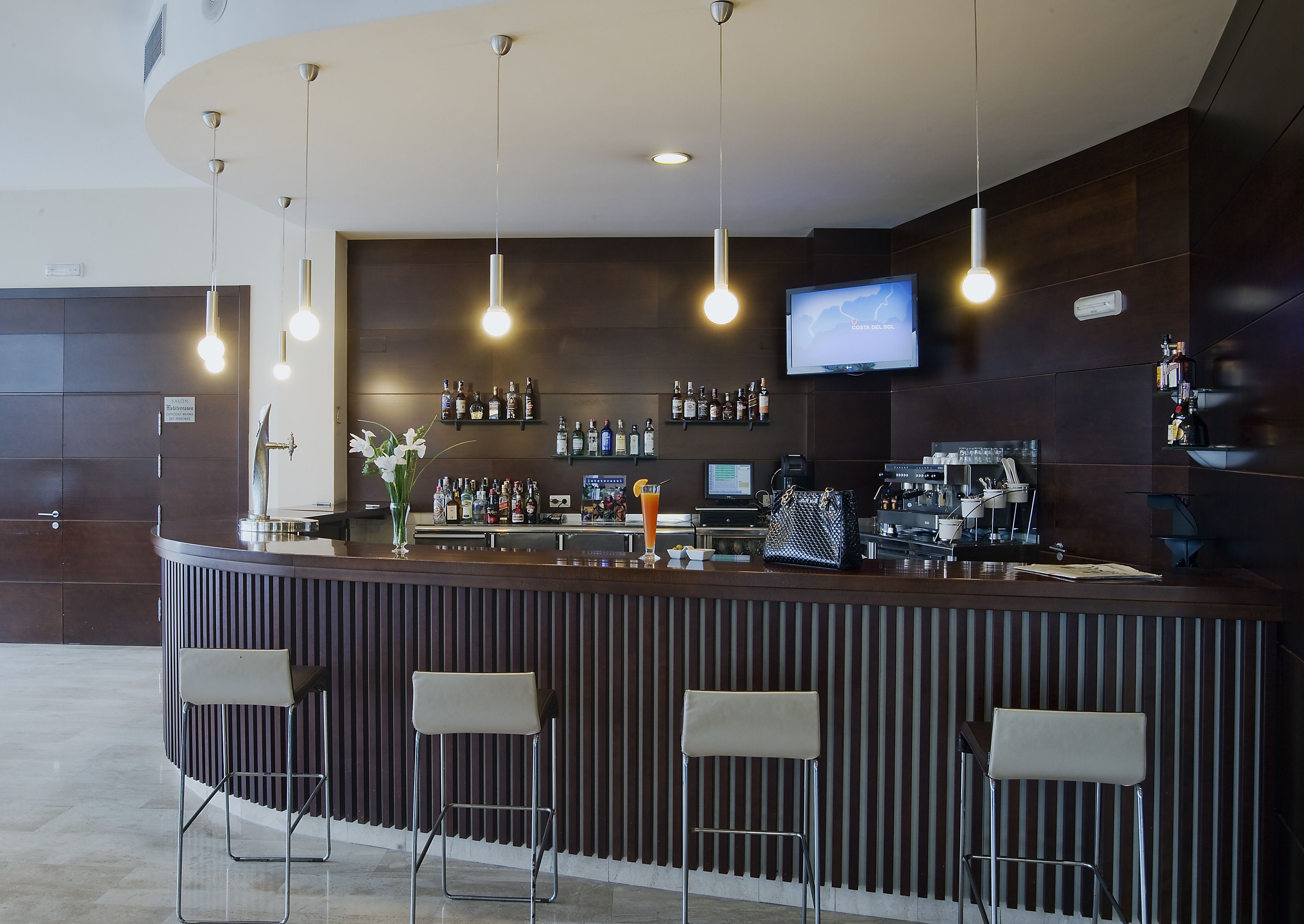 Angolo bar moderno con un bancone curvo, sgabelli da bar, luci a sospensione, un televisore e scaffali pieni di bottiglie.