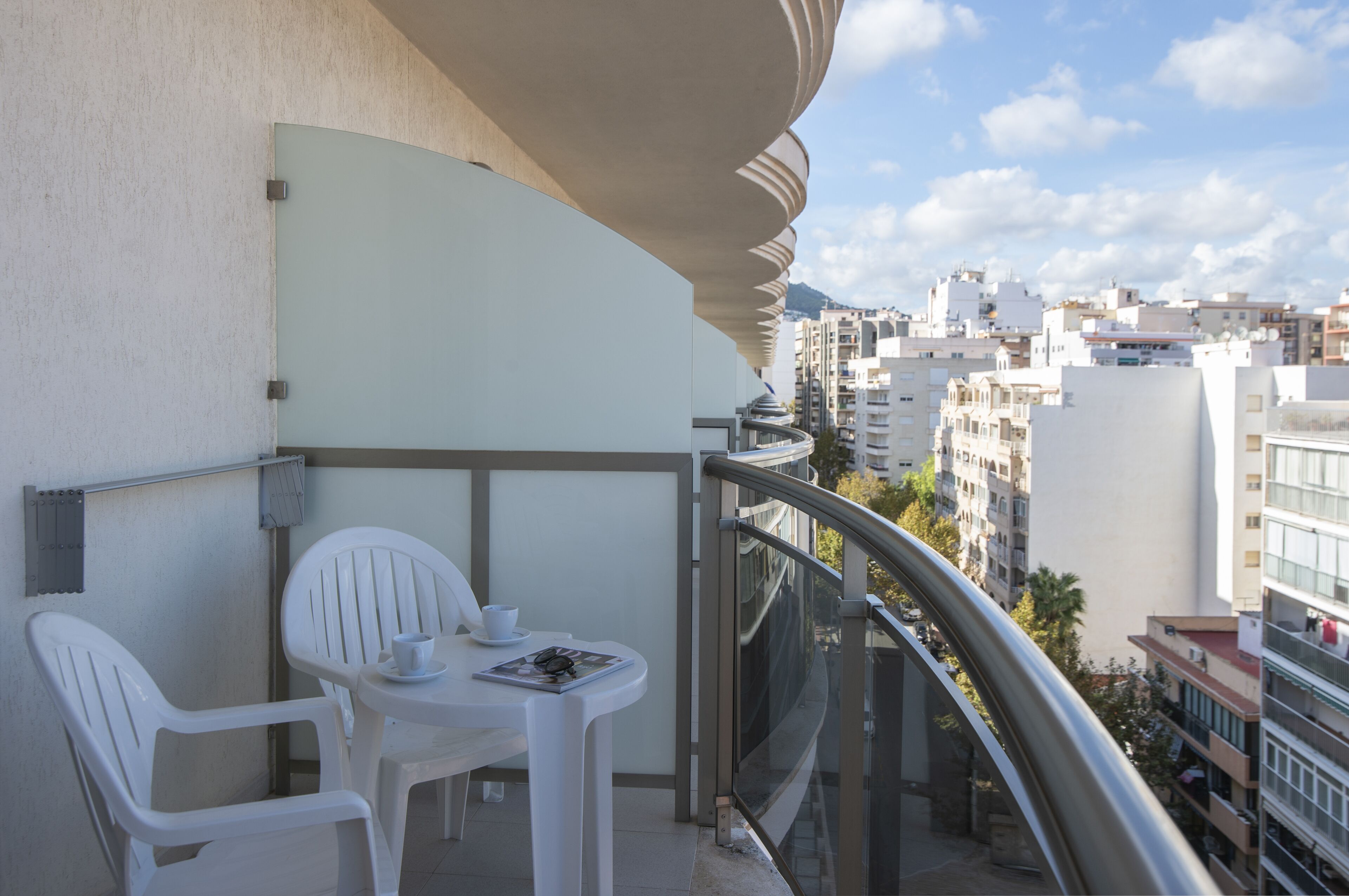 Een balkon met een witte tafel en stoelen, een glazen hekwerk en uitzicht op stadsgebouwen.