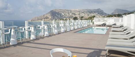 Uma área de refeições ao ar livre iluminada pelo sol, com piscina, cadeiras brancas e uma mesa posta com croissants, suco e um cardápio.