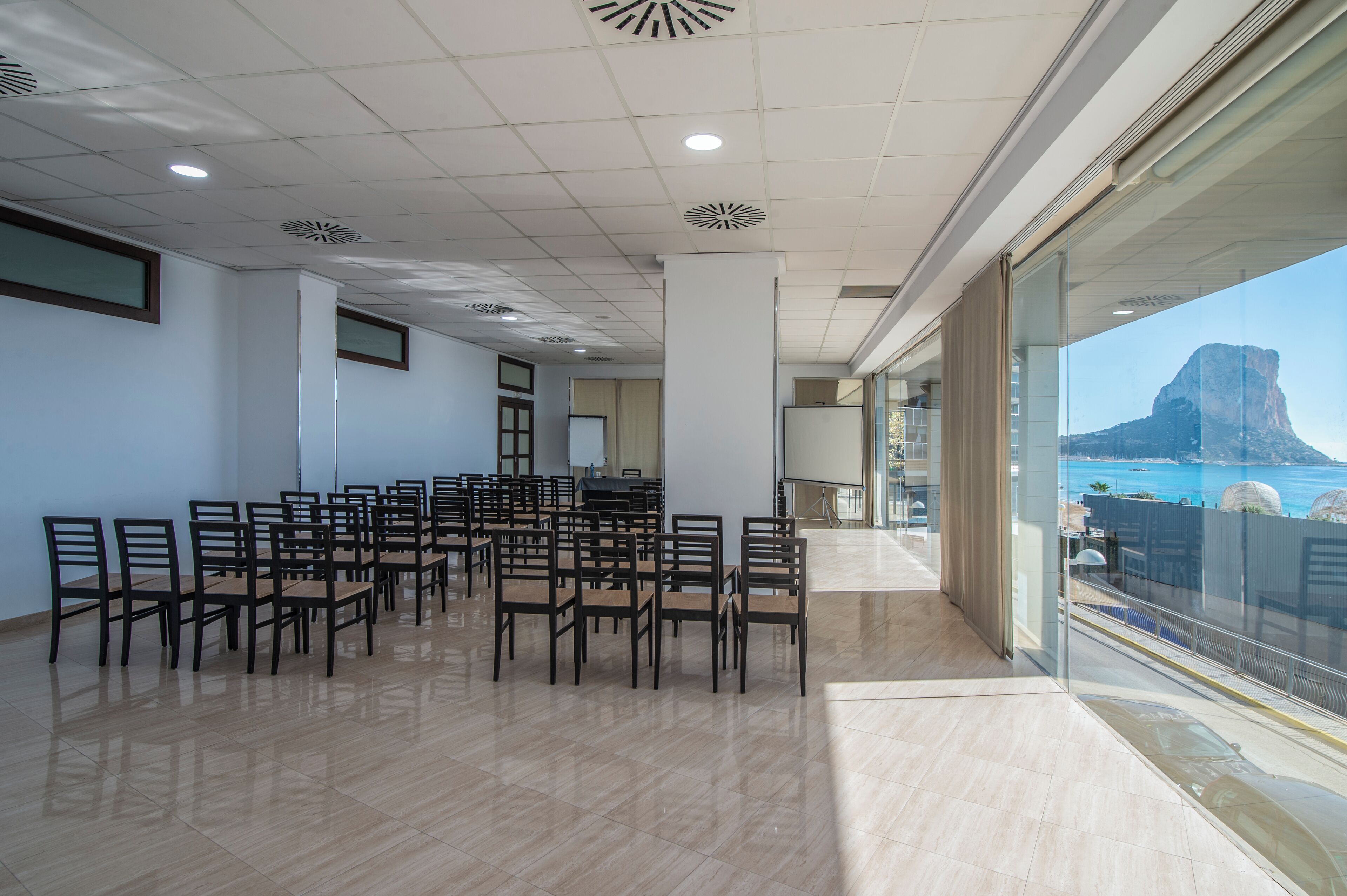 Ein Konferenzraum mit Stuhlreihen, einem großen Fenster mit Blick auf Berge und Meer sowie einer modernen Decke mit eingelassener Beleuchtung.