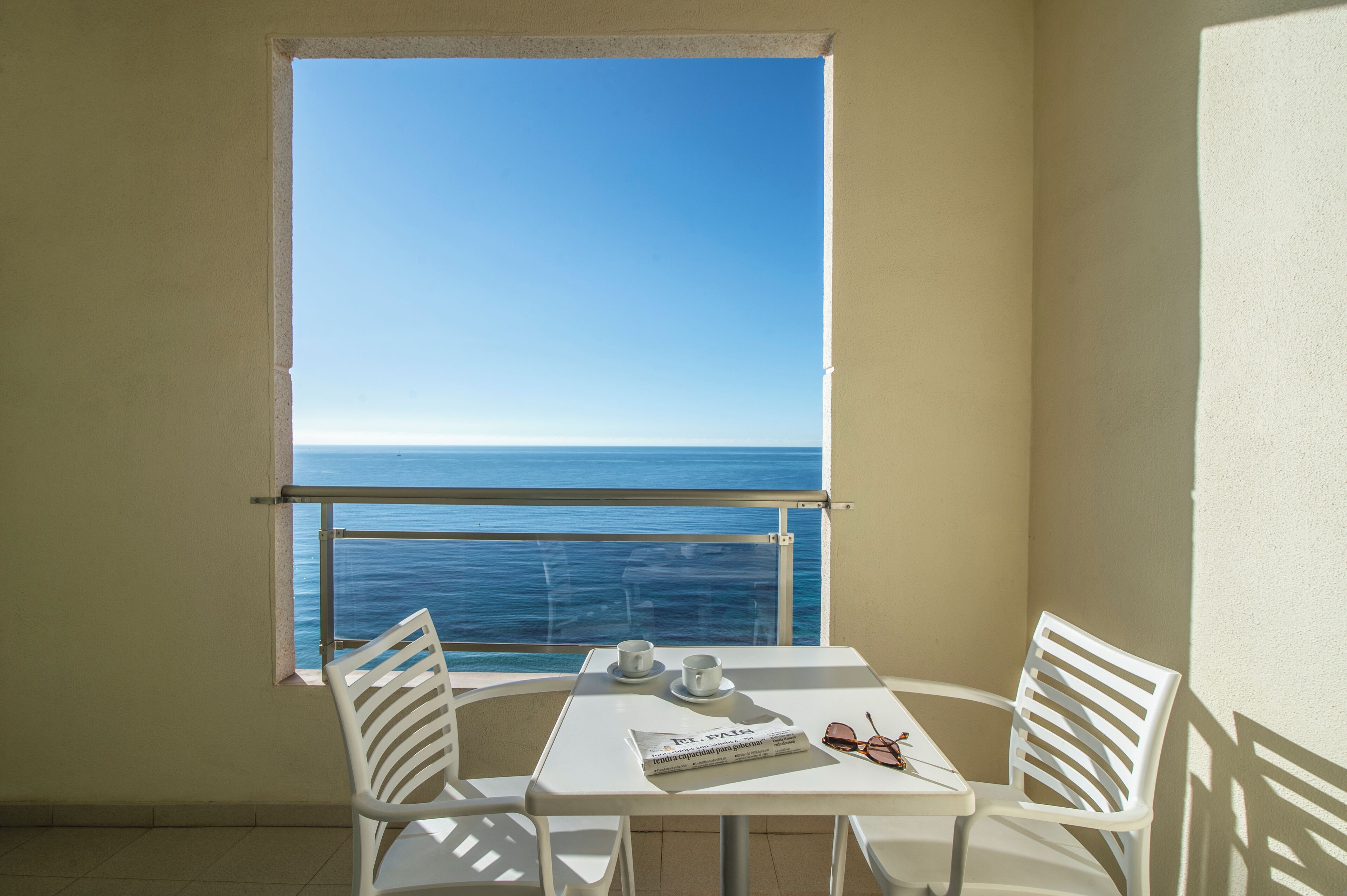 A balcony with a table and chairs, a window with a view of the sea, and a pair of sunglasses on the table.