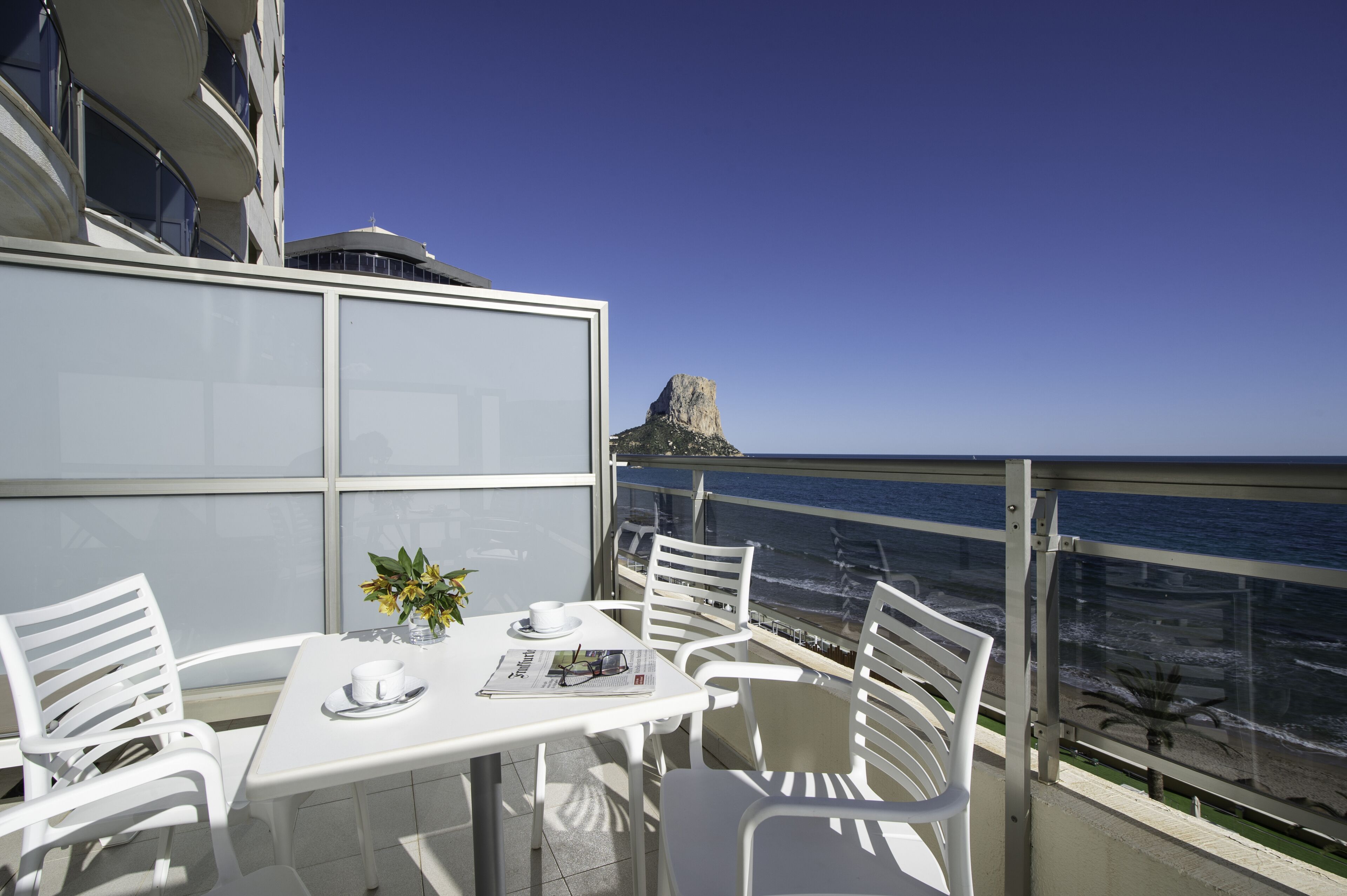 A balcony with white chairs and a table, overlooking the ocean with a rocky outcrop in the distance.